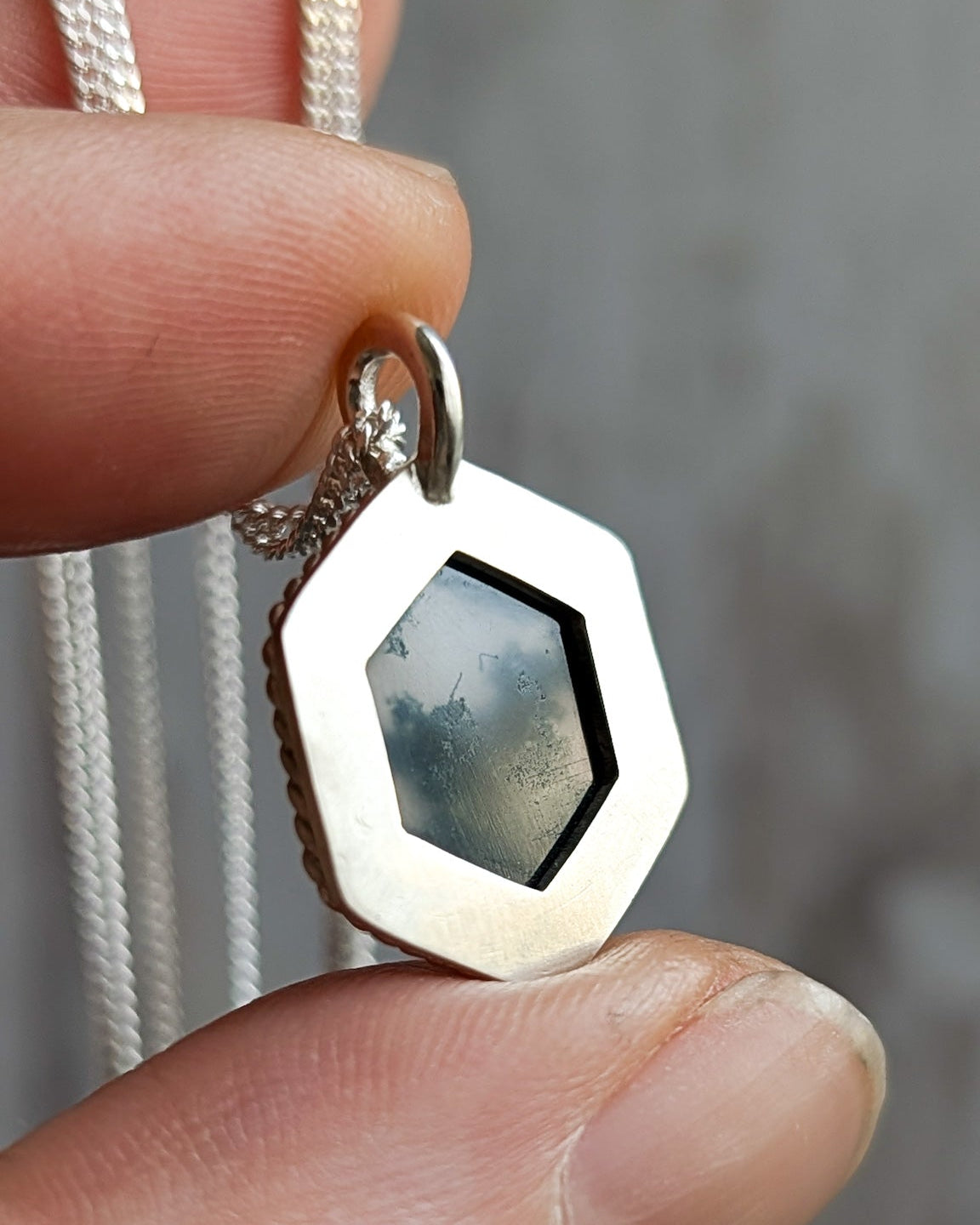 Closeup of a hand holding a silver pendant for size reference, showing the reverse side with a geometric shape cut-out revealing a mossy stone