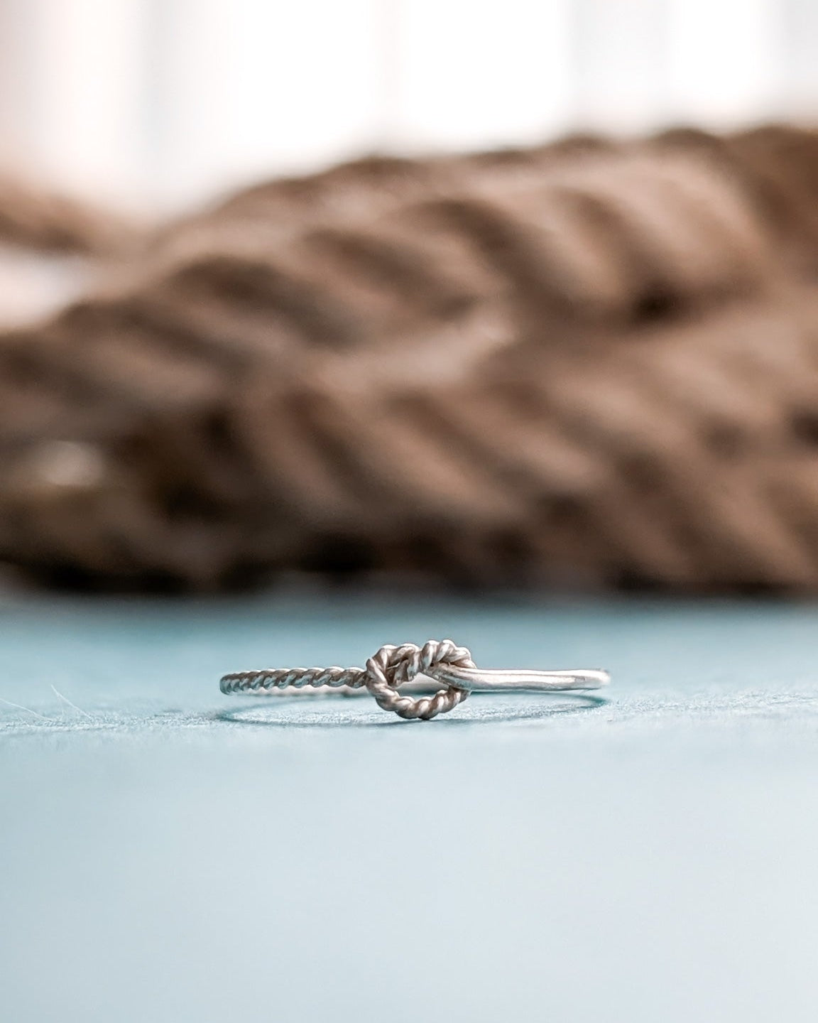 A silver ring with an overhand sailing knot detail, resting on a light blue surface with a blurred brown rope and soft background.