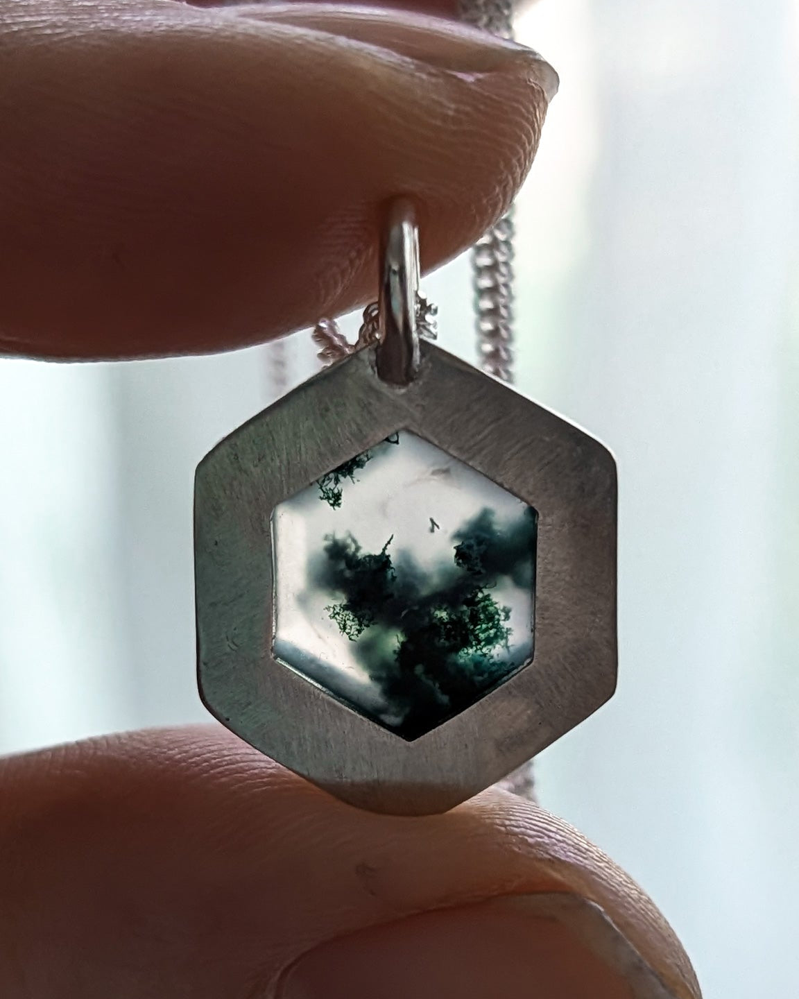 Hexagonal pendant with a scenic forest design held between fingers against a blurred bright background