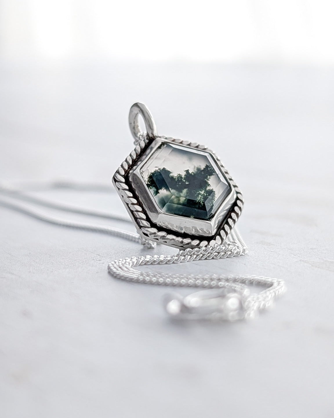 An off-centre view of a hexagon moss agate silver pendant on a bright white surface to highlight the green mossy texture inside