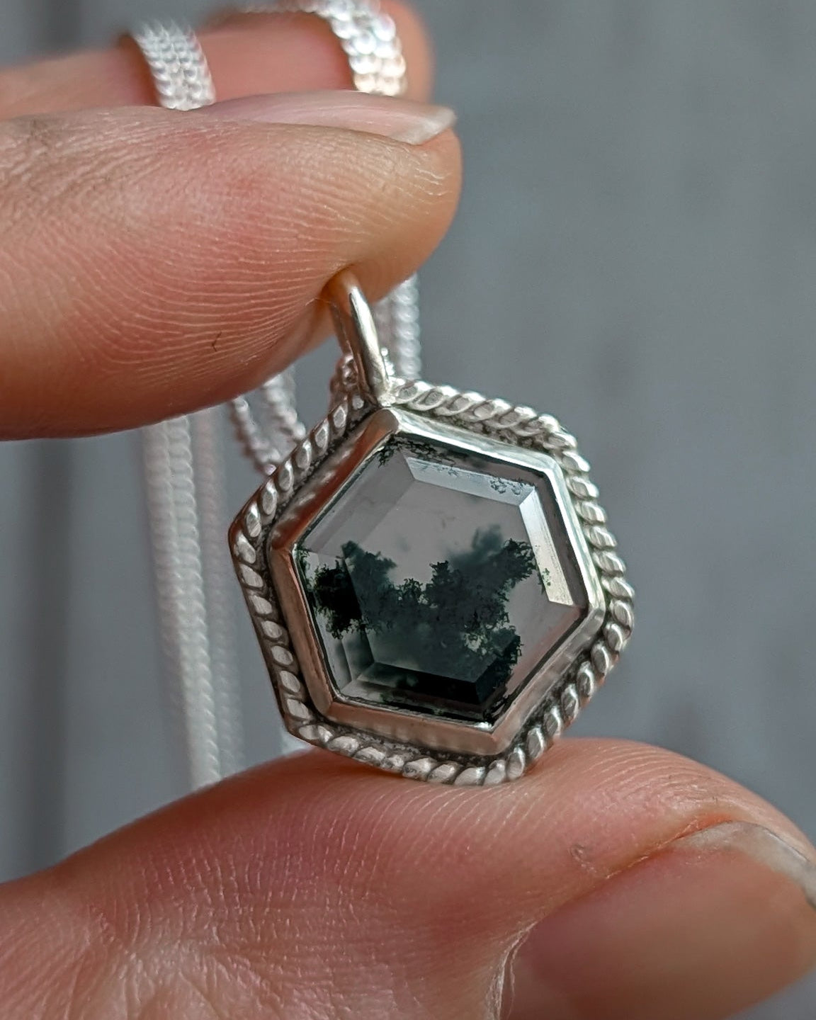 Silver hexagon moss agate pendant with a rope border, held between fingers with a blurred background
