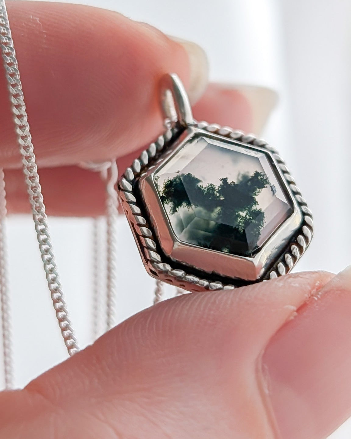 Close up of a hand holding a hexagonal silver pendant with mossy gemstone inside against a bright white background to highlight the moss-like green blooms inside