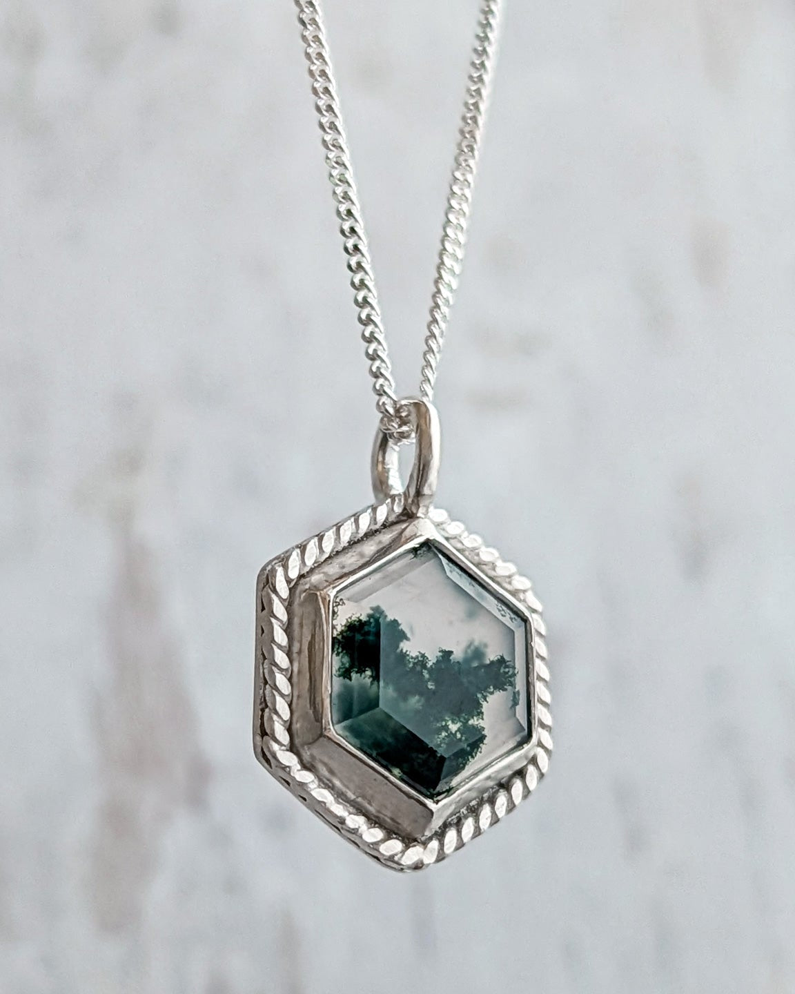Hexagonal silver pendant with a green mossy gemstone hanging in front of a white textured background