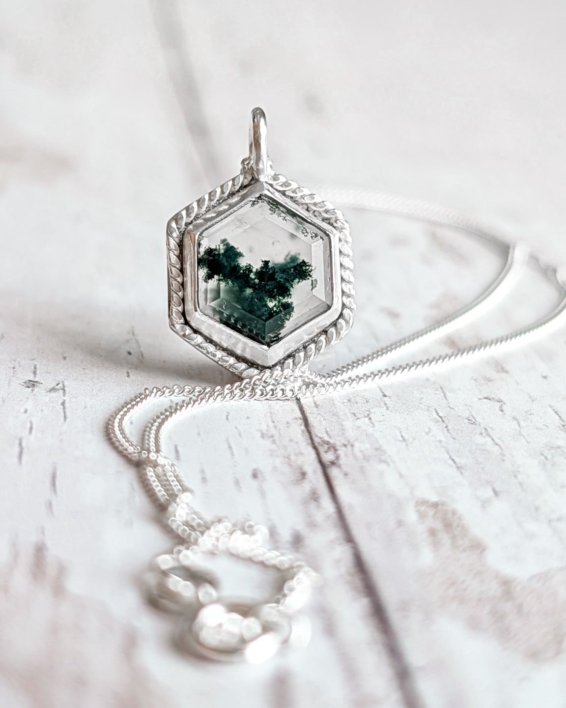 Silver hexagon pendant with green moss gemstone on a white wooden surface