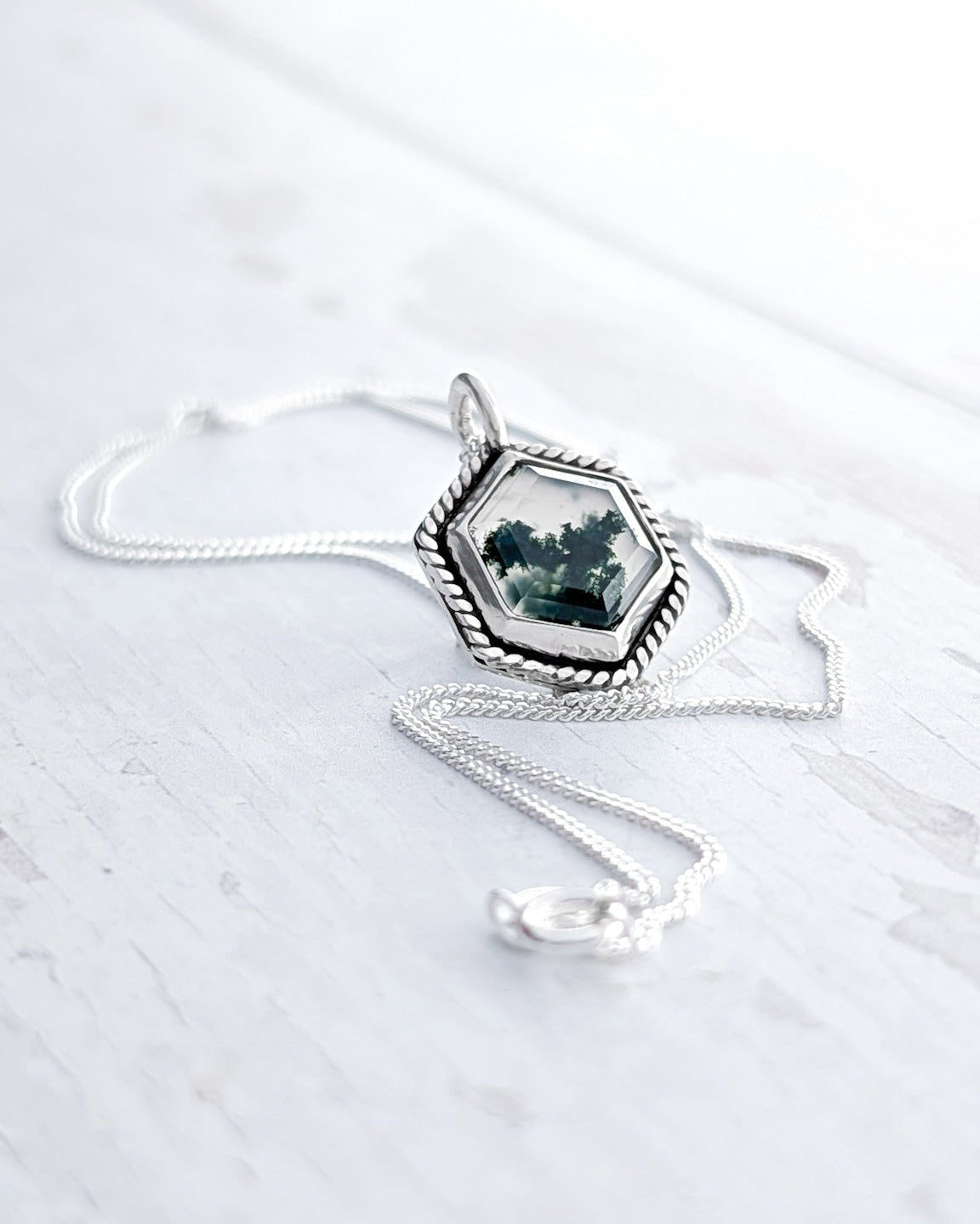 A silver pendant charm featuring a hexagon moss agate stone rests amongst a silver chain on a bright white textured surface