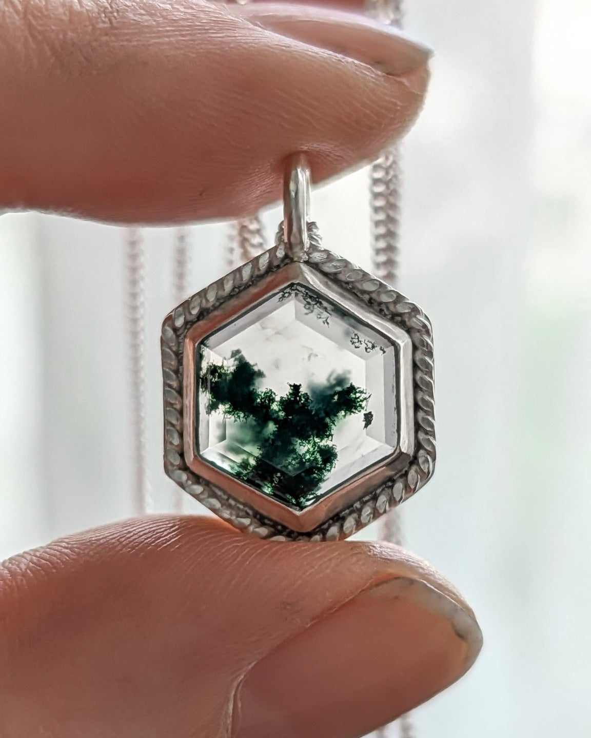Closeup of a hexagonal pendant with green mossy stone held between fingers against a blurred white background