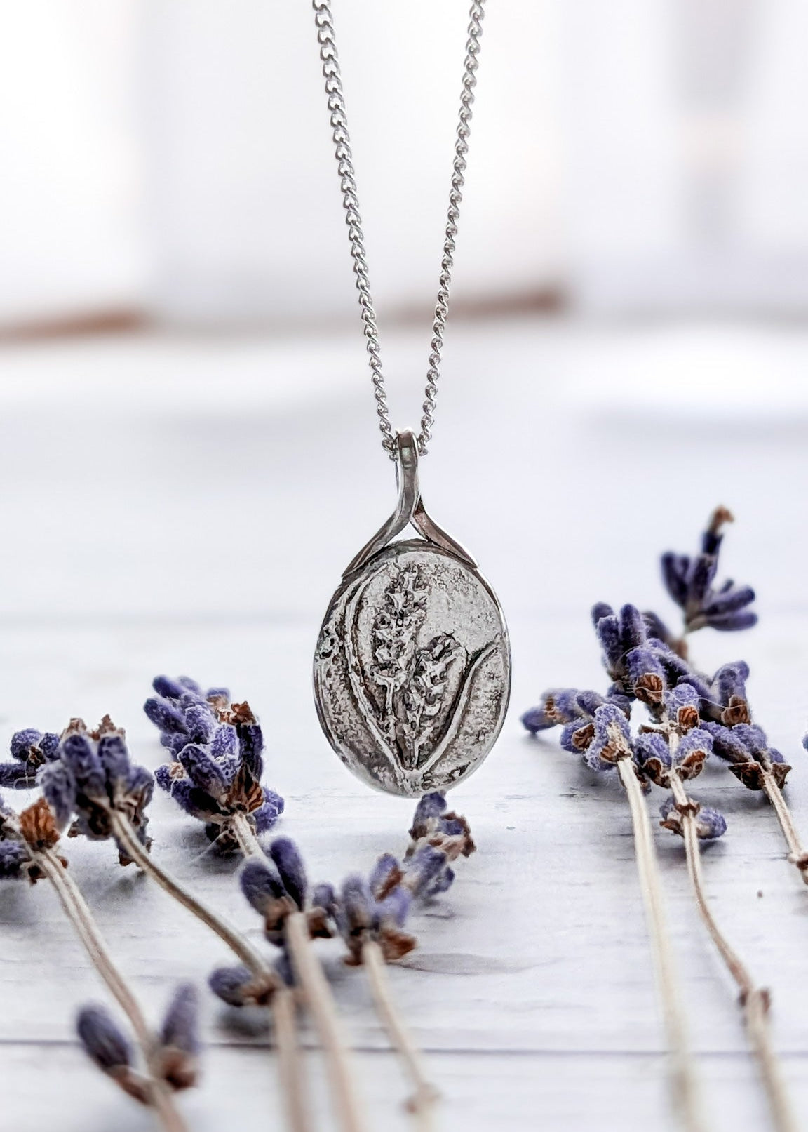 A small silver oval pendant with floral details embossed on the front alongside organic texture, pictured amongst scattered dried lavender on a white wooden surface and gentle white fabric background