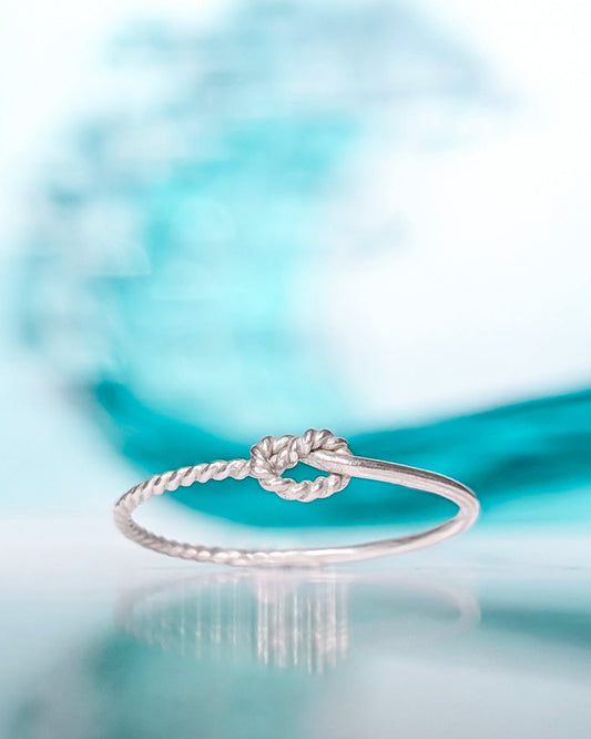 A delicate silver rope-knot ring resting on a reflective surface with a soft, blurred turquoise wave and white background.