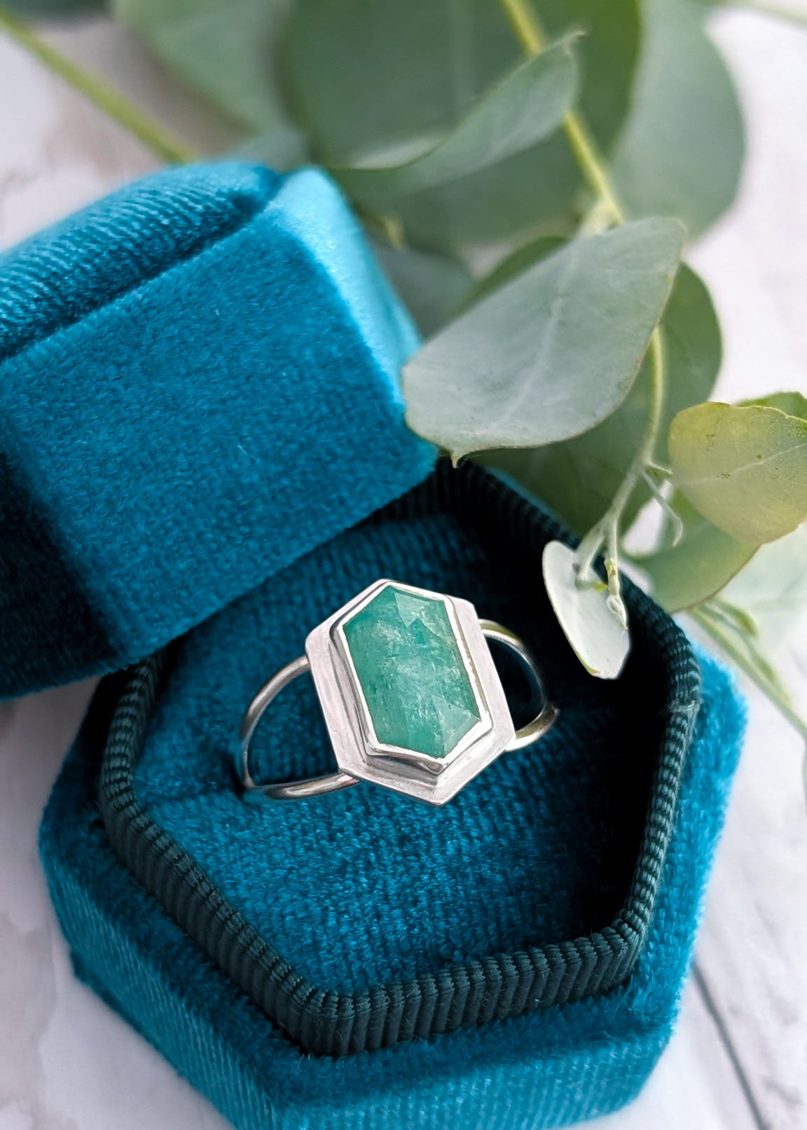 A blue velvet hexagon ring box sits open with a pale green gemstone ring sitting inside, backed by some decorative plants