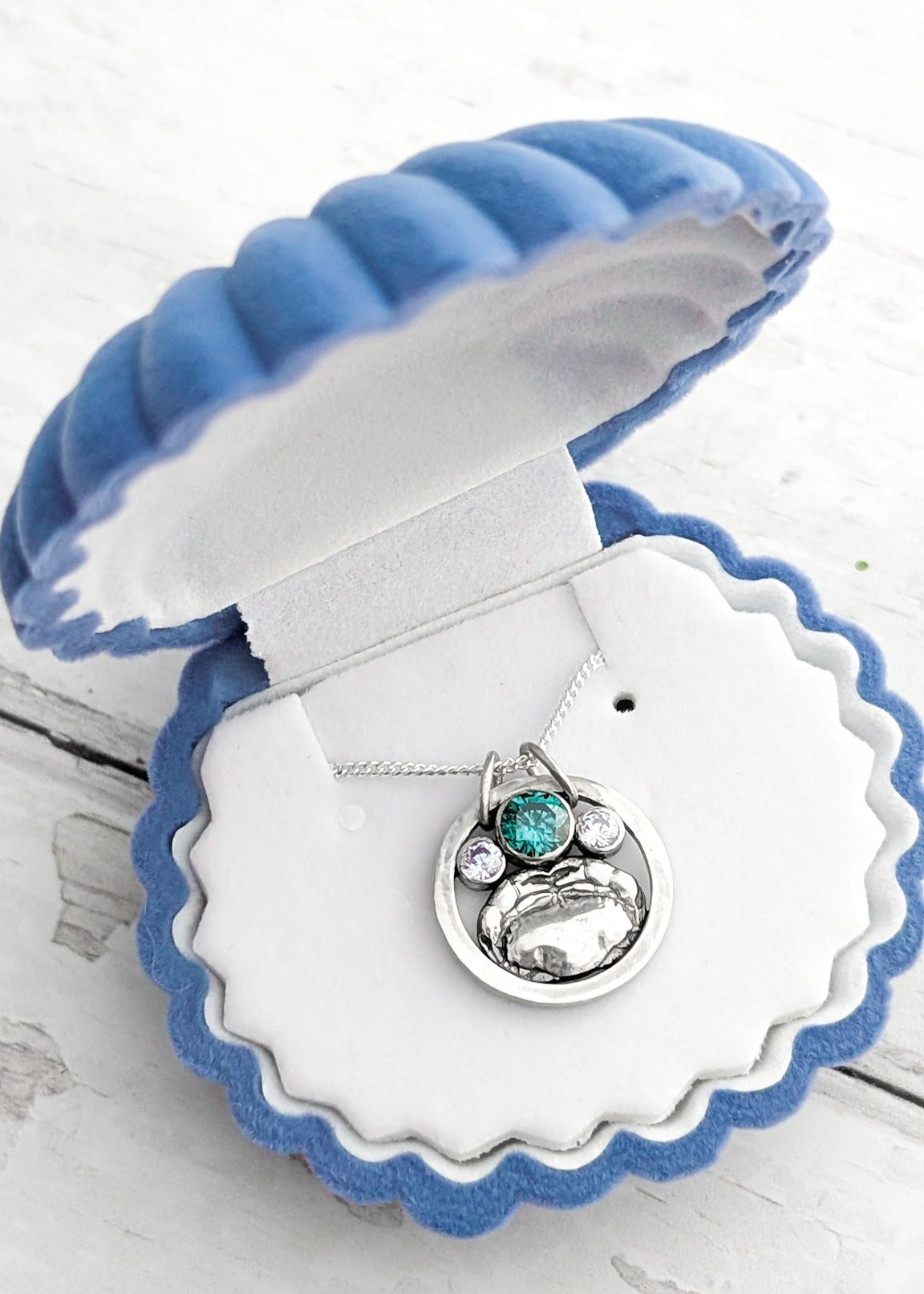 Round silver necklace featuring a crab and sparkling round gemstones in a blue seashell jewelry box on a light wooden surface.