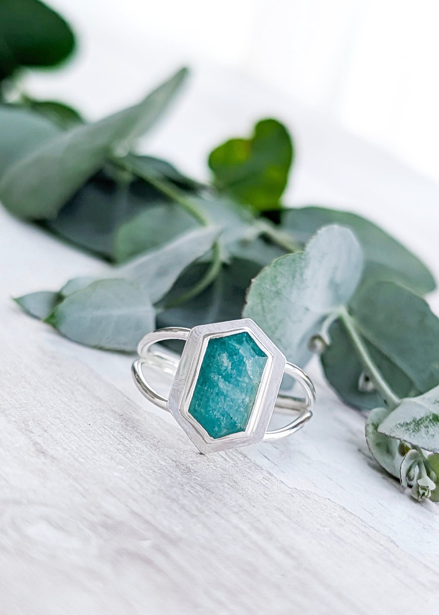 A pale turquoise Amazonite hexagon ring rests on a white wooden surface in front of some soft focus leaves