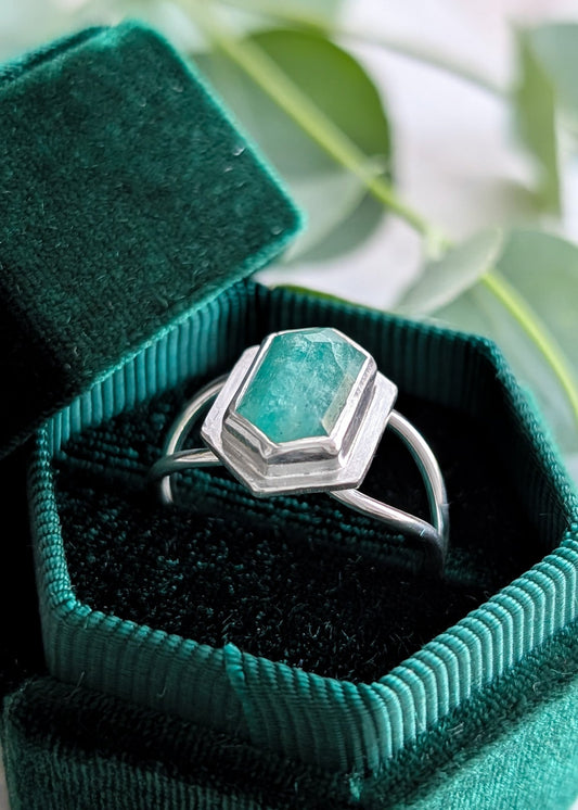 Closeup of a hexagonal velvet ring box with a silver ring inside, the ring has a large pale green geometric cut gemstone in the centre