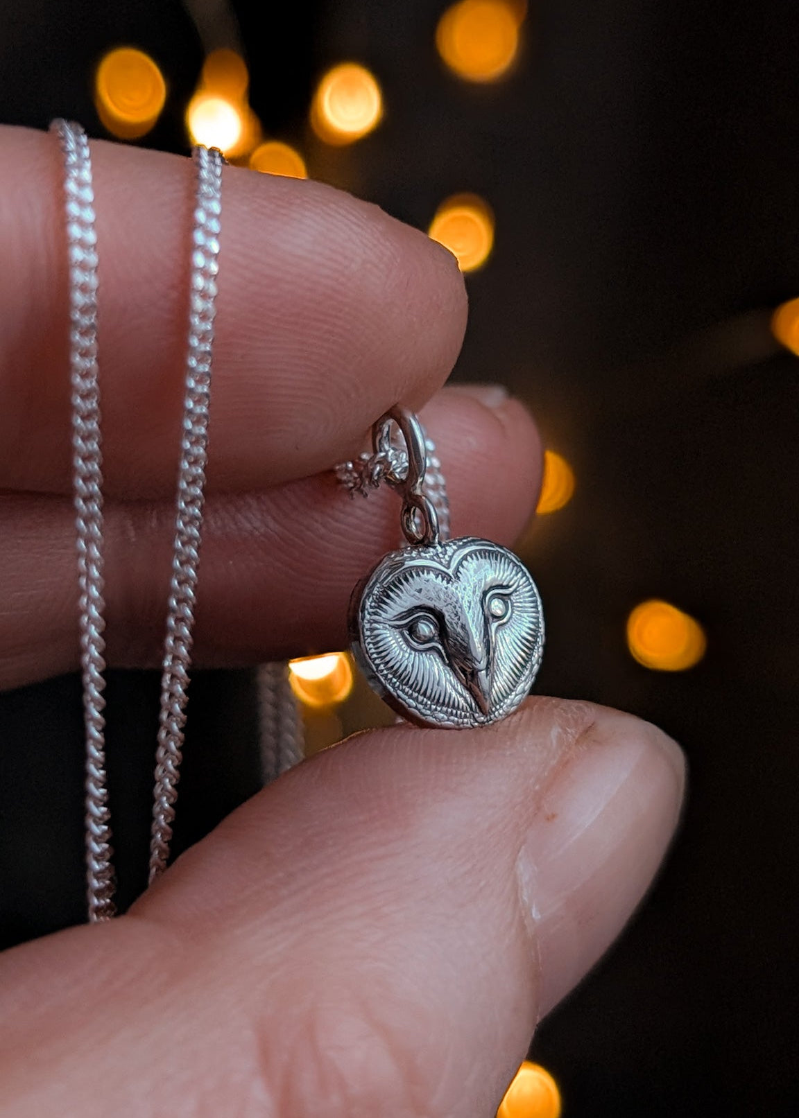 Silver necklace with detailed barn owl face charm held between fingers against a blurred bokeh lights background