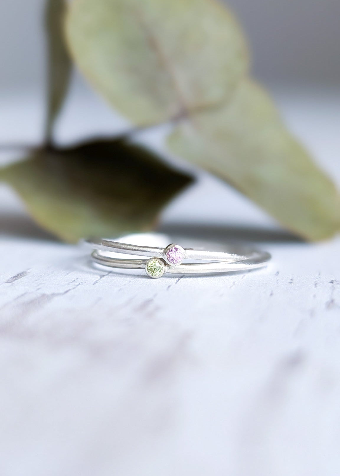 Pair of dainty silver stacking rings, one with a pastel pink stone and one with a pale green stone.  Pictured stacked together on a white wooden surface in front of some soft focus leaves.
