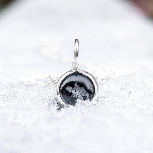Small silver pendant featuring a black stone with a real preserved snowflake in the centre, photographed on a blanket of fresh snow.
