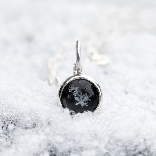 Silver pendant featuring a round black stone with some small genuine preserved snowflakes in the centre, photographed resting on a blanket of fresh snow
