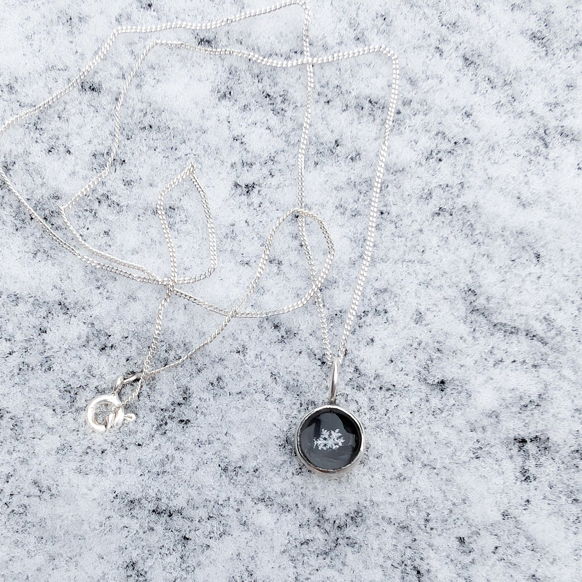 A circular black charm on a silver necklace in the snow, the black circle features a genuine white snowflake in the middle.