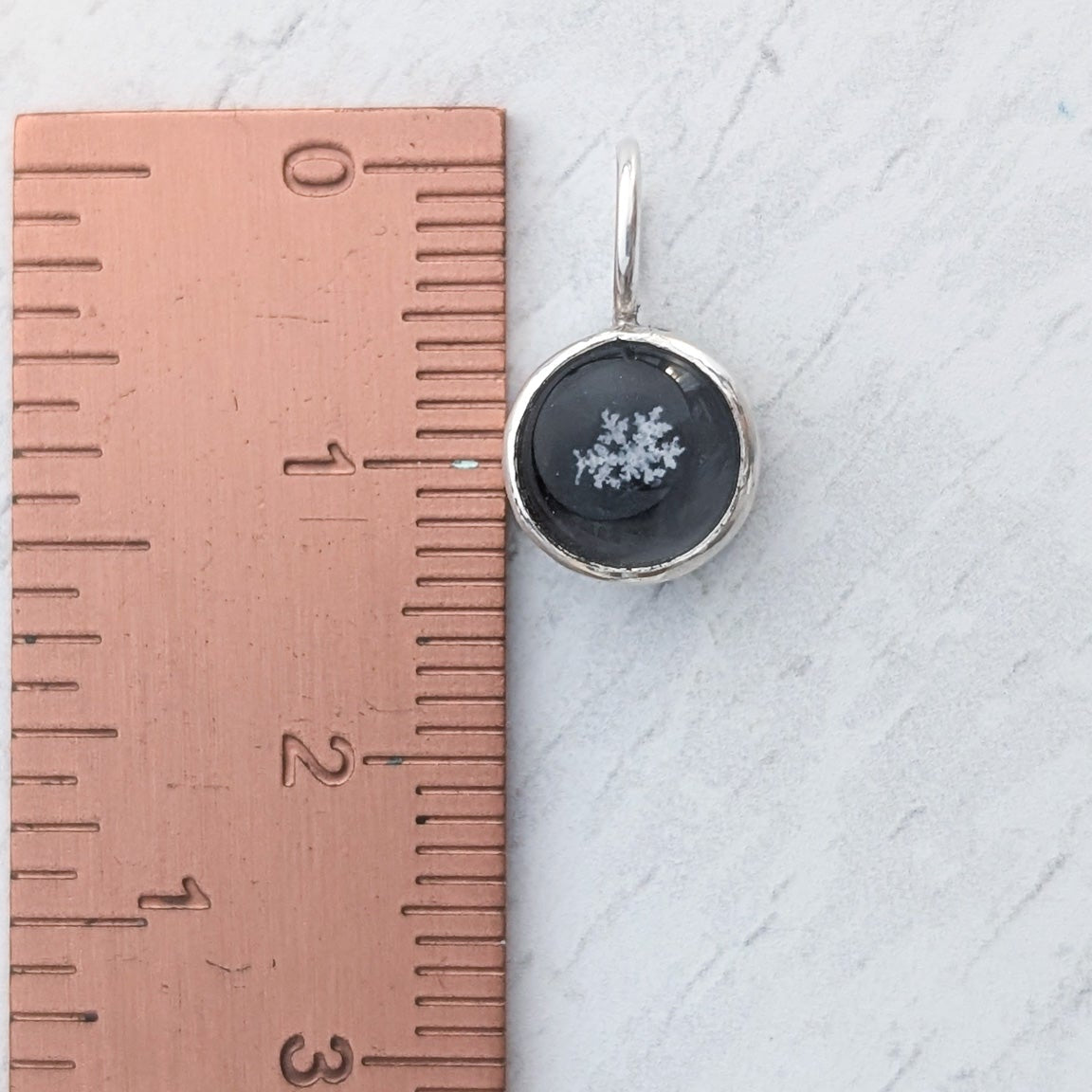 Small round pendant with a genuine snowflake preserved in the centre, pictured on a light wooden background beside a ruler for scale.