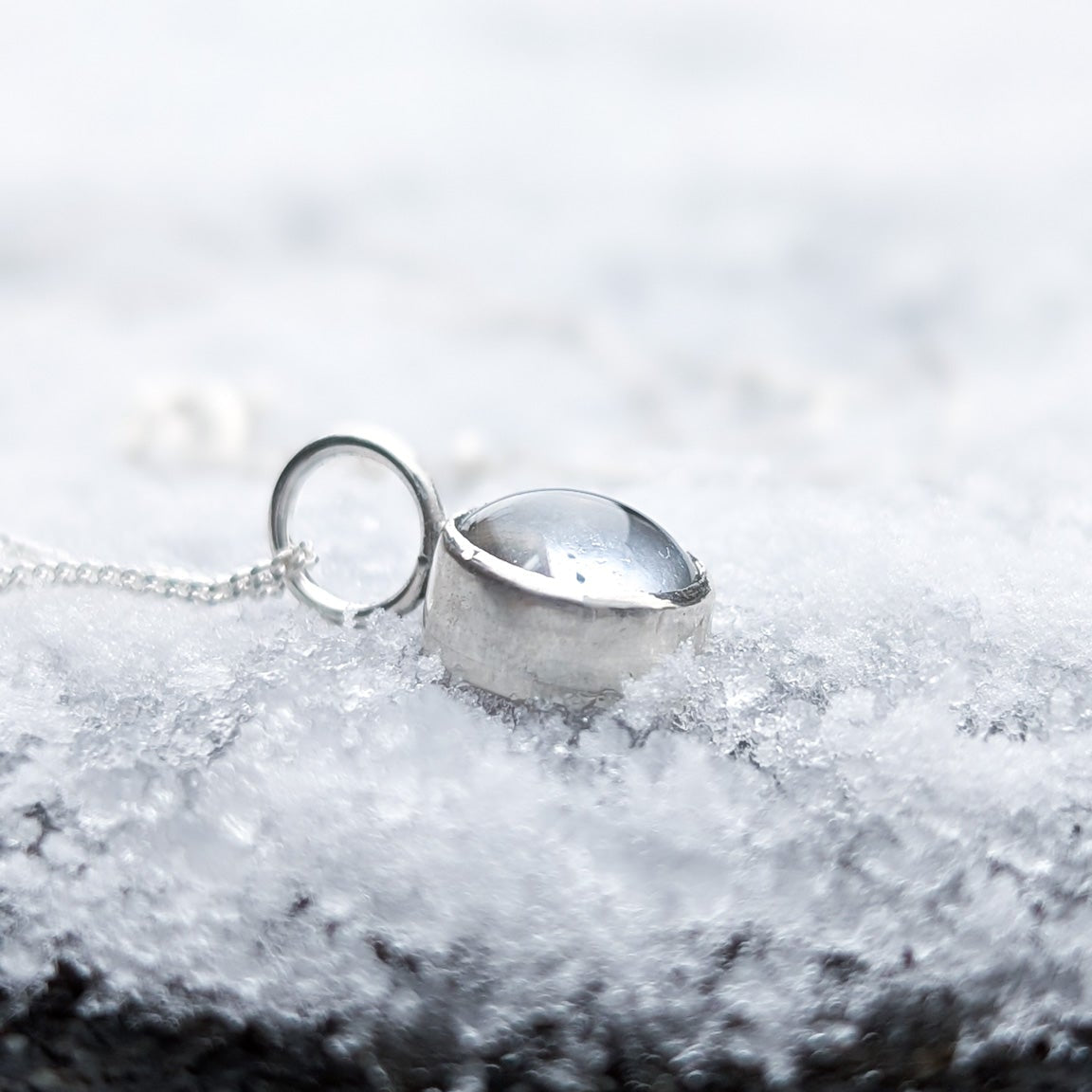 Side view of a small silver pendant hanging from a large jumpring and chain, holding a domed glass cabochon.  The necklace is resting on fresh snow.