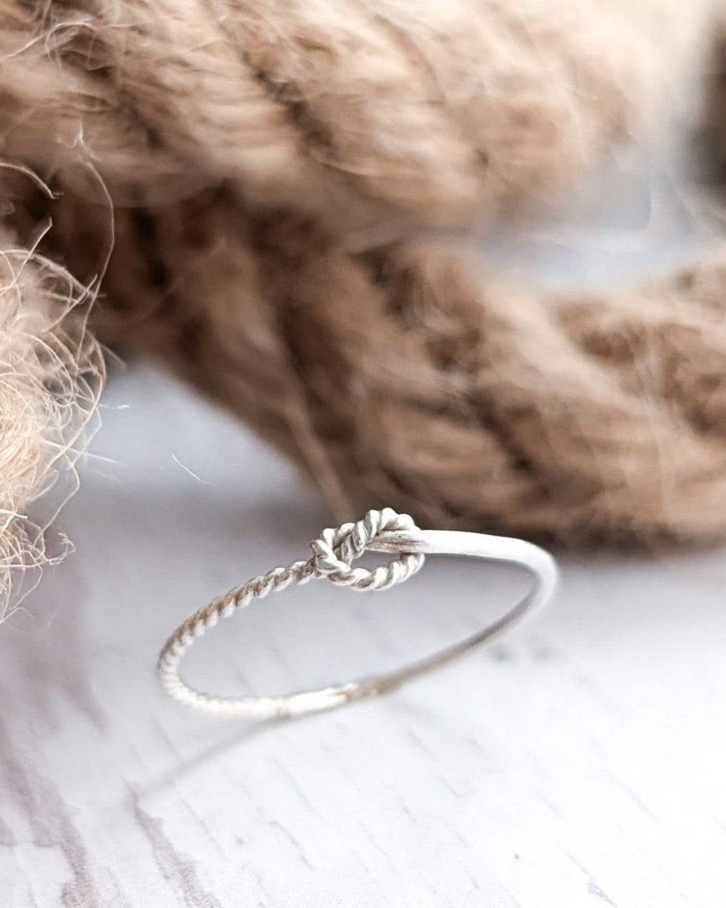 A dainty silver ring with a nautical knot rope design, resting on a white wooden surface with a soft-focus background of sailing rope.