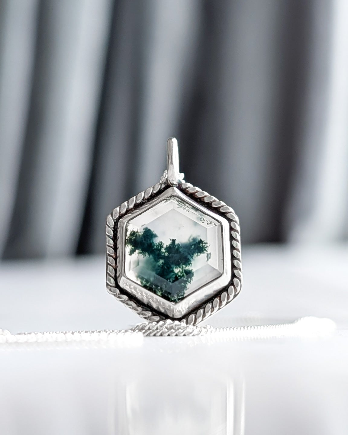 Closeup of a hexagonal moss agate stone set in a silver necklace on a reflective white surface in front of a textured grey background