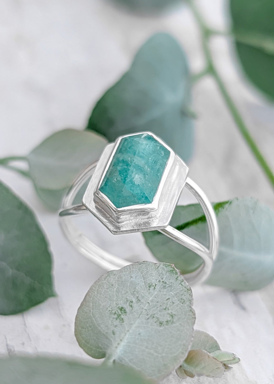 Close up of a double banded silver ring with oblong hexagon pale teal gemstone, the ring sits amongst soft green eucalyptus leaves