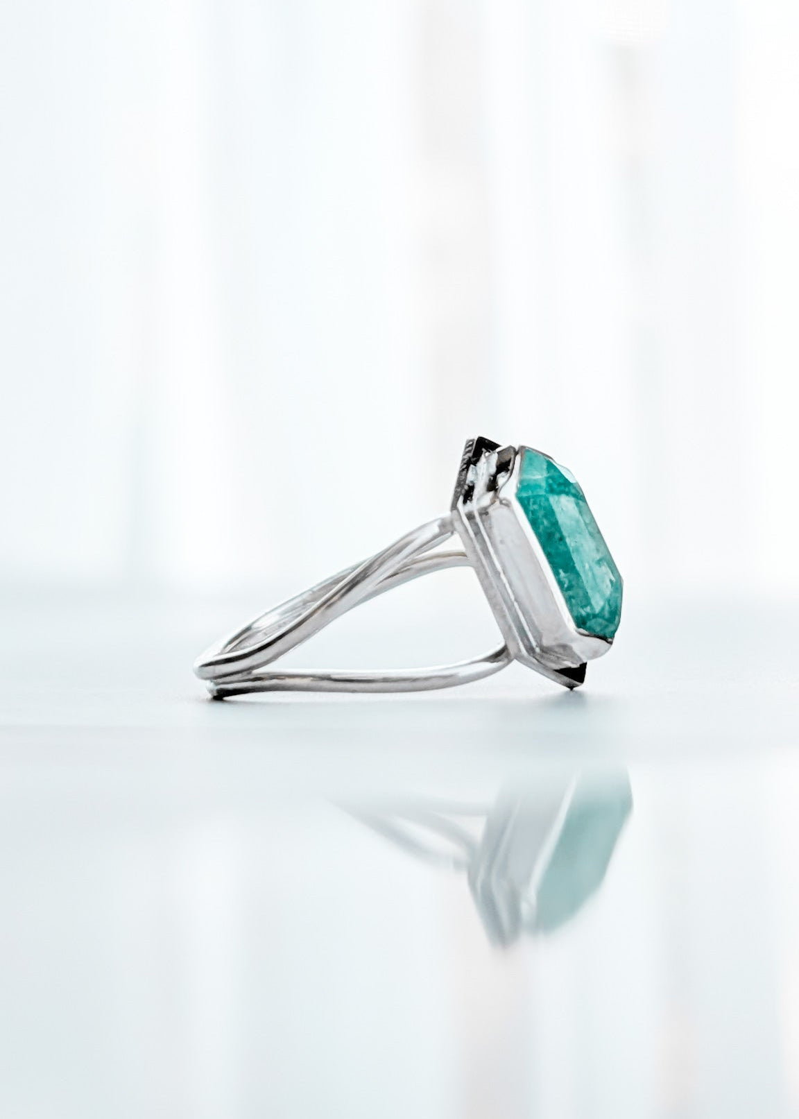 Side view of a silver ring featuring a 1cm cushion-cut emerald green gemstone, photographed on a reflective white surface with a soft, bright background.