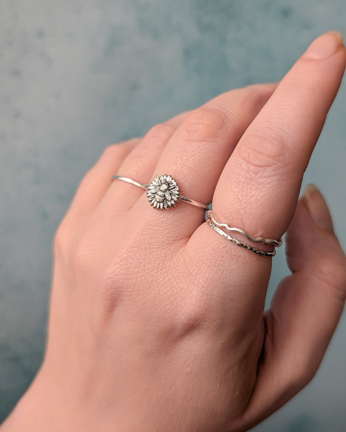 Hand wearing several silver rings, the middle finger displays a silver bee and flower design, blurred blue background