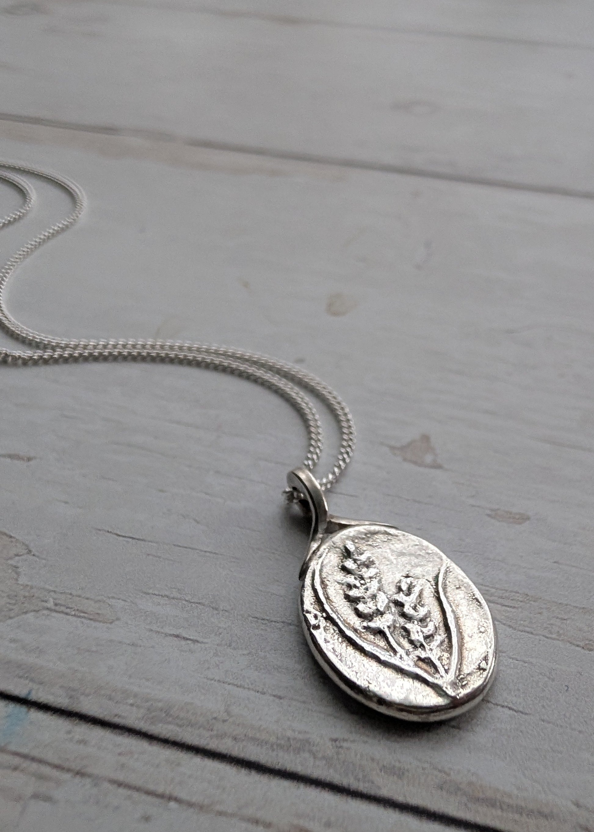 An organically textured silver oval pendant with botanical and floral detail embossed on the front, resting with its silver chain snaking behind it on a light wooden surface