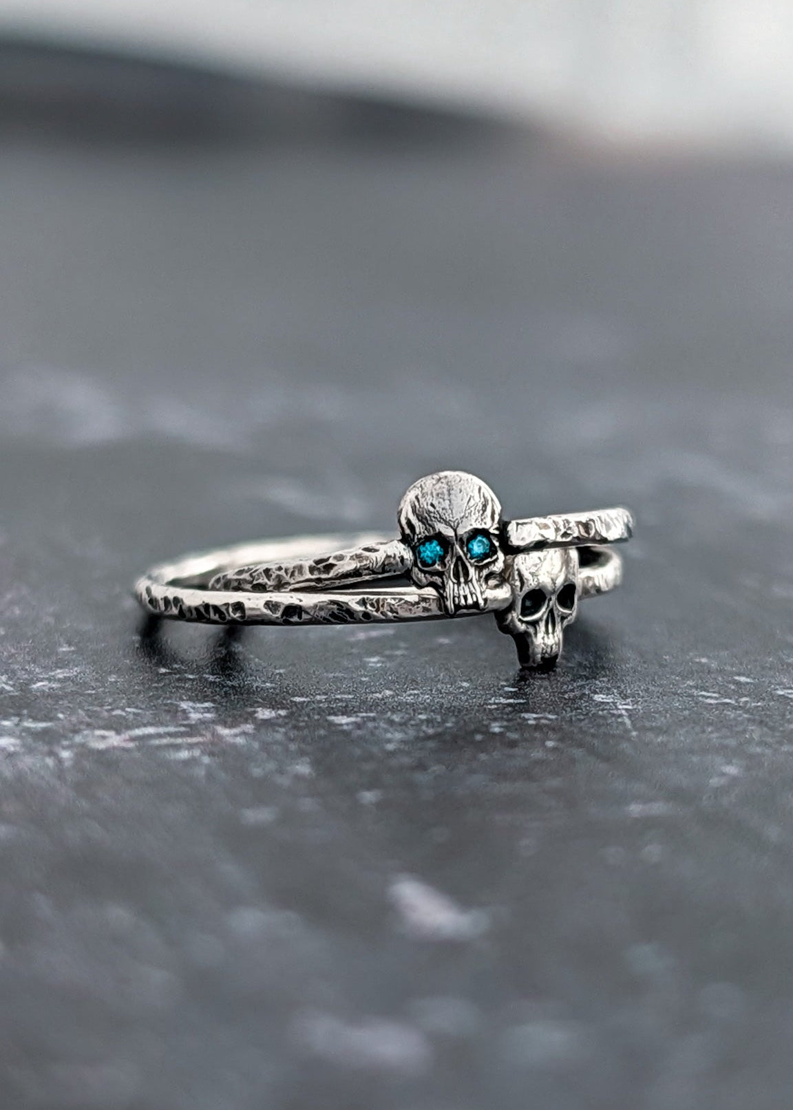 A silver skull ring with small blue gemstone eyes, resting on a dark textured surface.
