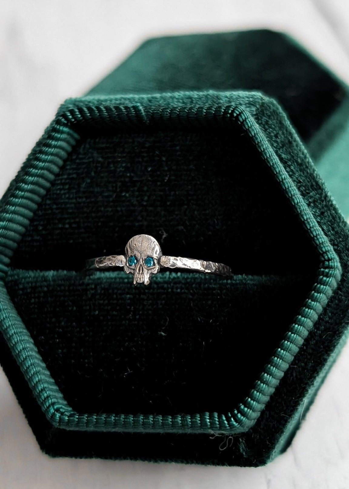 A silver skull ring with small blue gemstone eyes displayed in an open dark green velvet hexagonal ring box on a white marble surface.