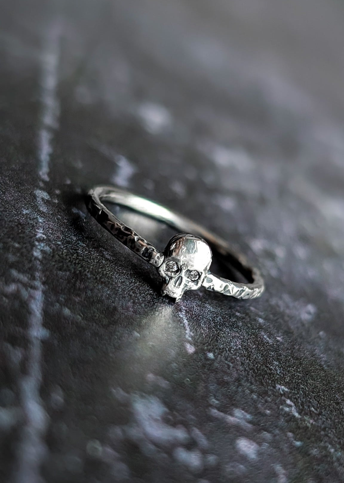 A silver skull ring with clear gemstones set in the eyes rests on a dark, textured grey surface.