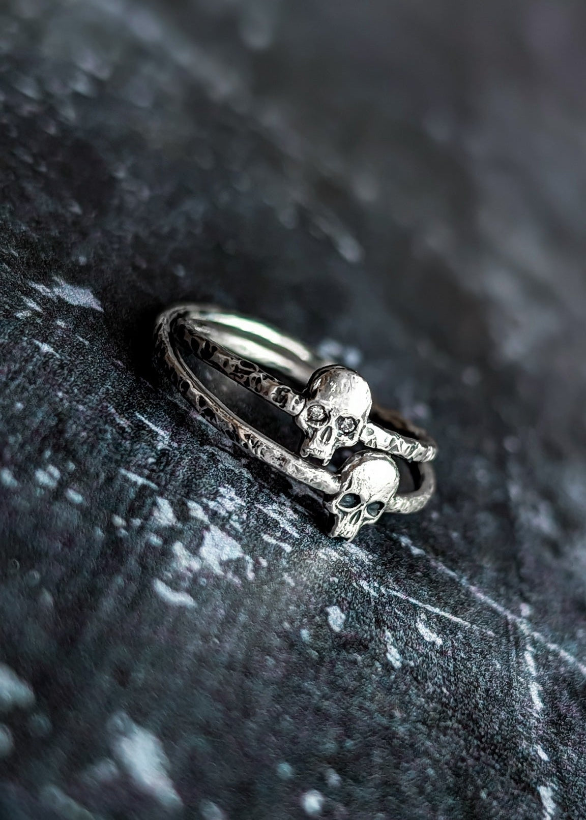 Two silver gothic rings featuring small skull motifs and textured band details, rest on top of each other on a dark, textured stone surface.