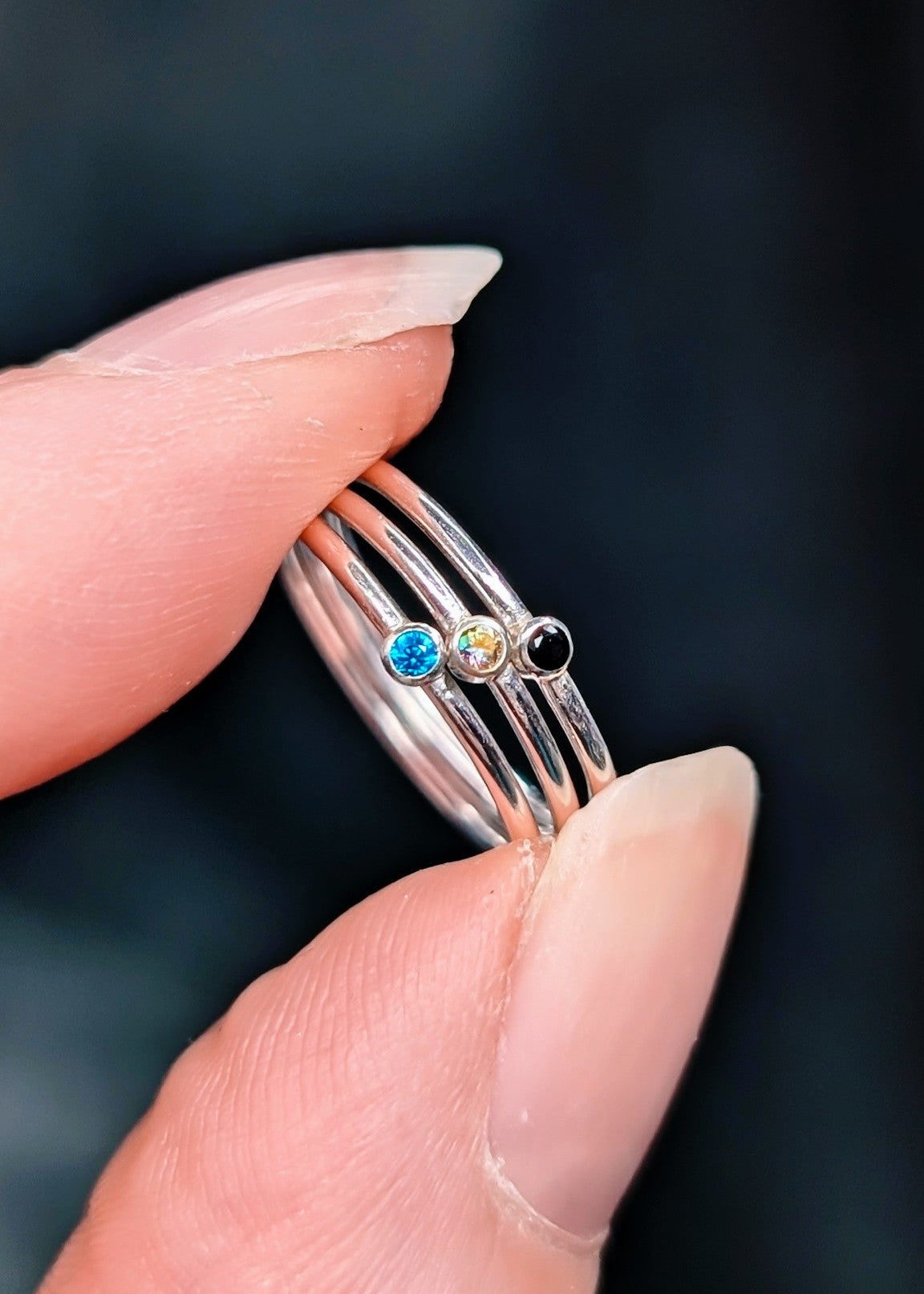 Close up of an index finger and thumb holding a stack of 3 sparkling stone rings, each a different colour.  Pictured on a dark background.