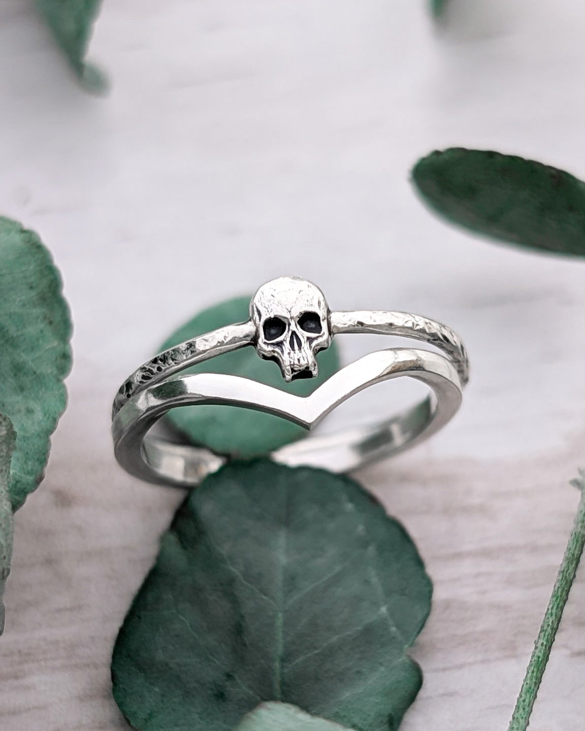 A silver wishbone ring stacked with a silver skull ring, photographed with scattered leaves on a white wooden surface