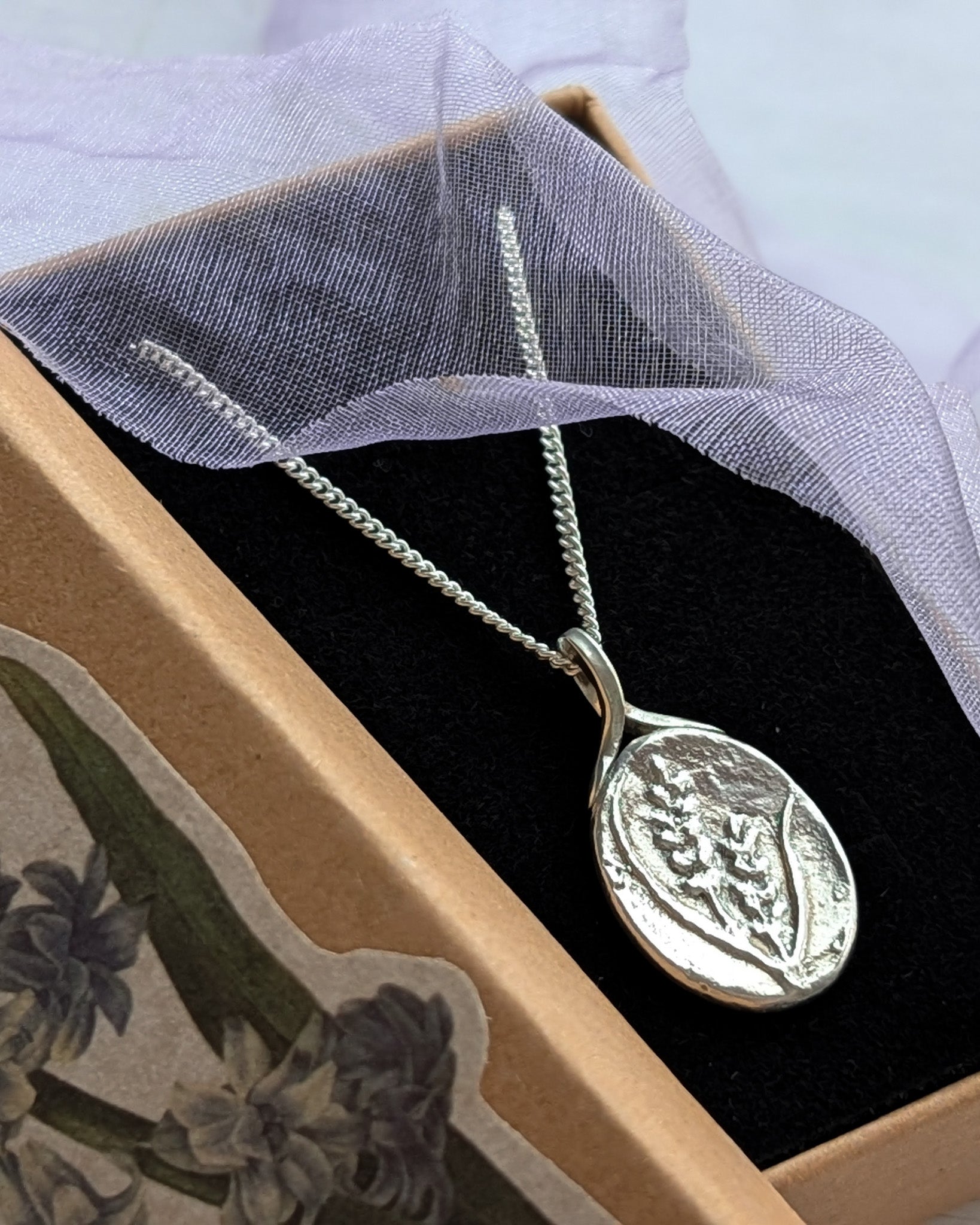 A silver oval pendant with embossed lavender flowers and grasses on the face, pictured in an open jewellery gift box with some lavender ribbon