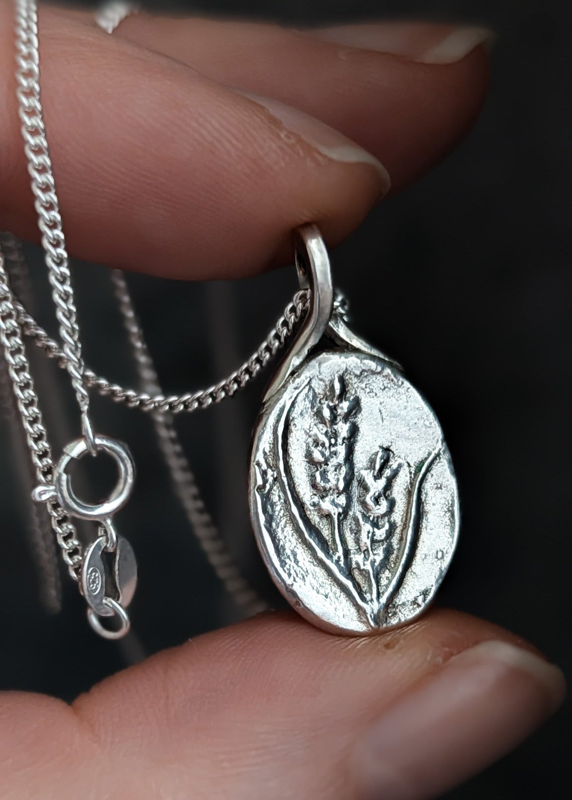 Closeup of a hand holding an oval floral pendant between the thumb and index finger against a dark background that highlights the shine of the silver