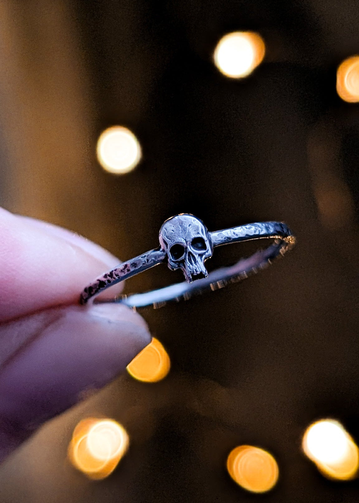 Skinny ring band with central skull charm held by a hand with blurred lights in the background