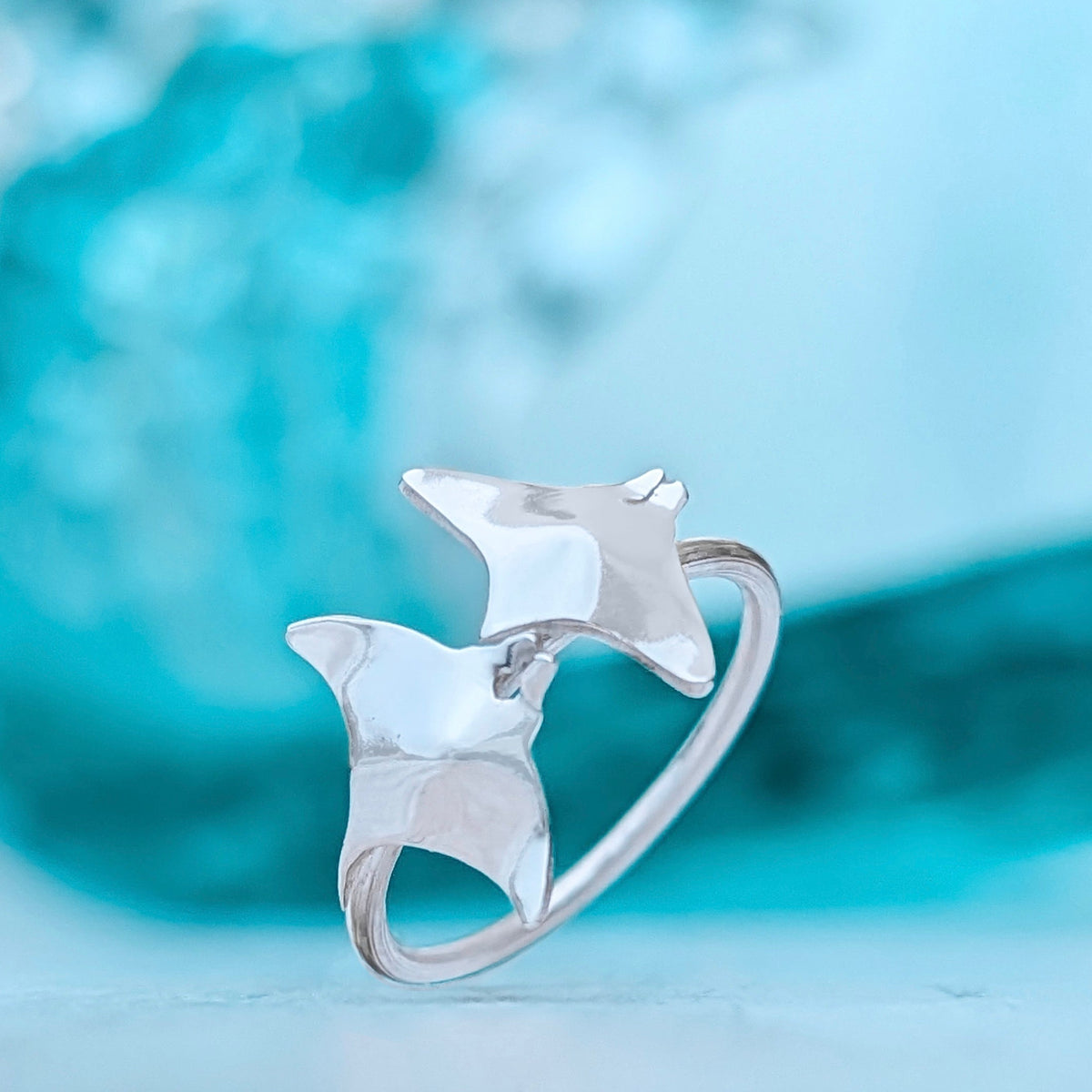 A shiny silver ring with two 3D manta shapes on it rests suspended at an angle in front of a blurred focus wave background