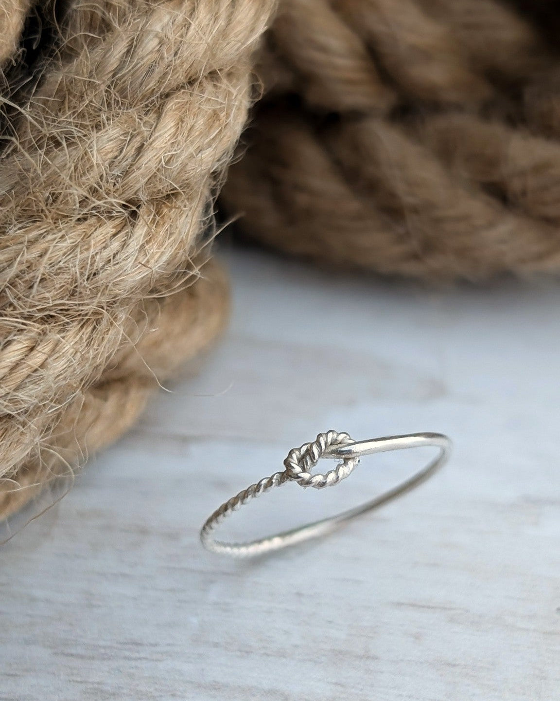 A petite silver ring with knotted rope detail, displayed on a light wooden surface with a blurred background of natural jute rope.