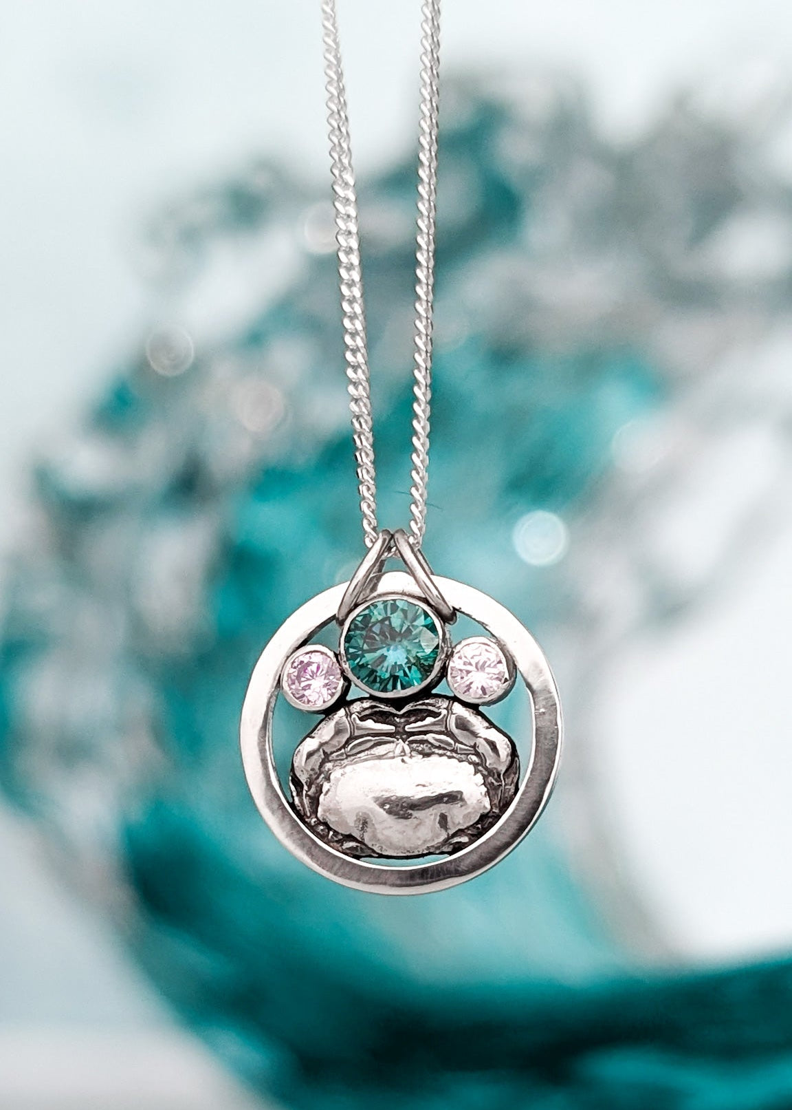 Silver circle and crab pendant necklace with teal green gemstone and smaller pale pink stones on a blurred teal ocean background