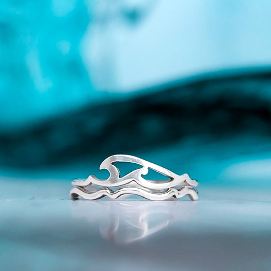 A silver tribal wave ring stacked on top of a crimped wavy silver ring band, pictured in front of a blue cresting ocean wave