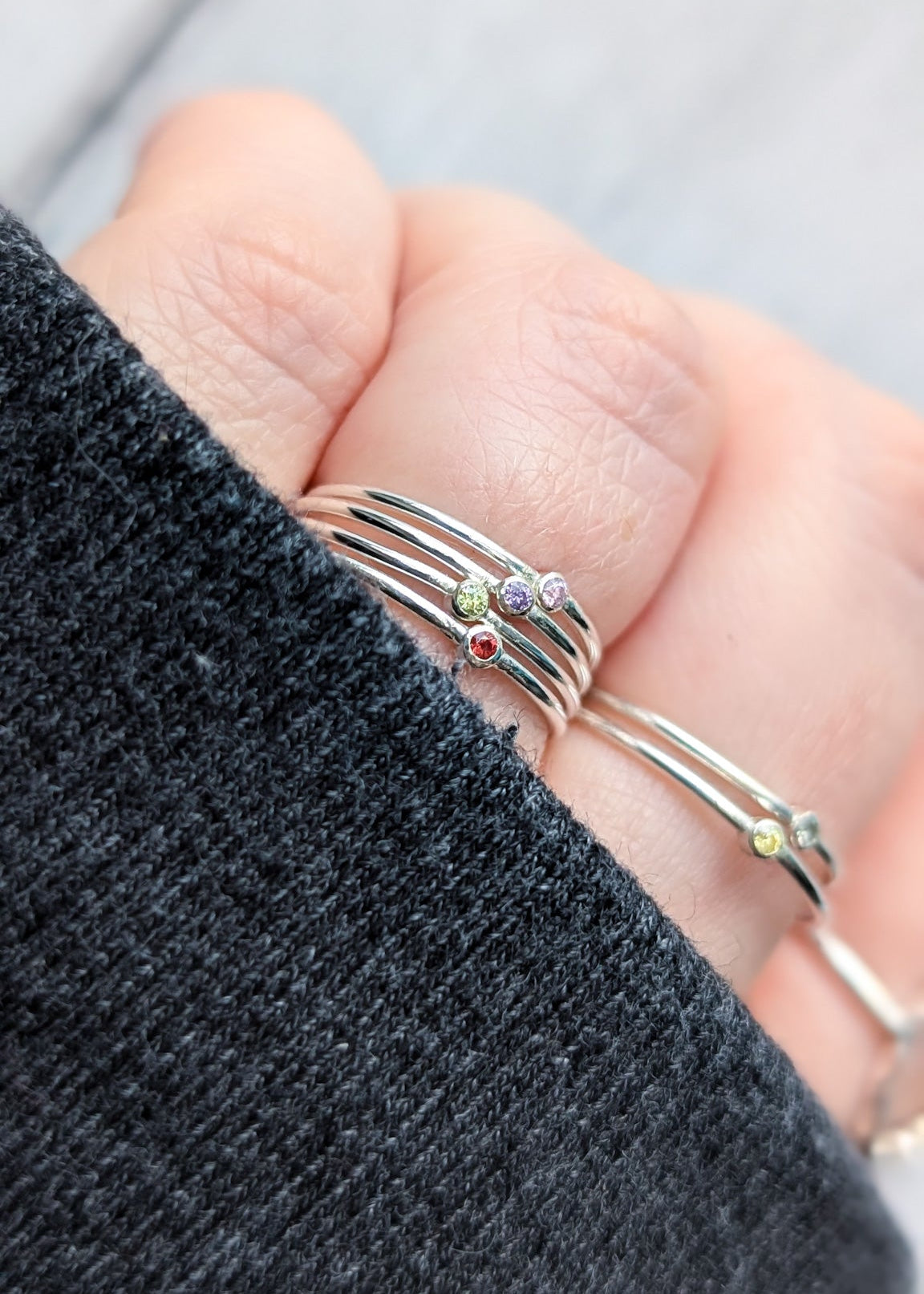 Close up of a hand, partially covered by a knitted sleeve, with fingers all wearing petite silver stacking rings.  Each ring has one tiny stone set in the centre, all different colours.