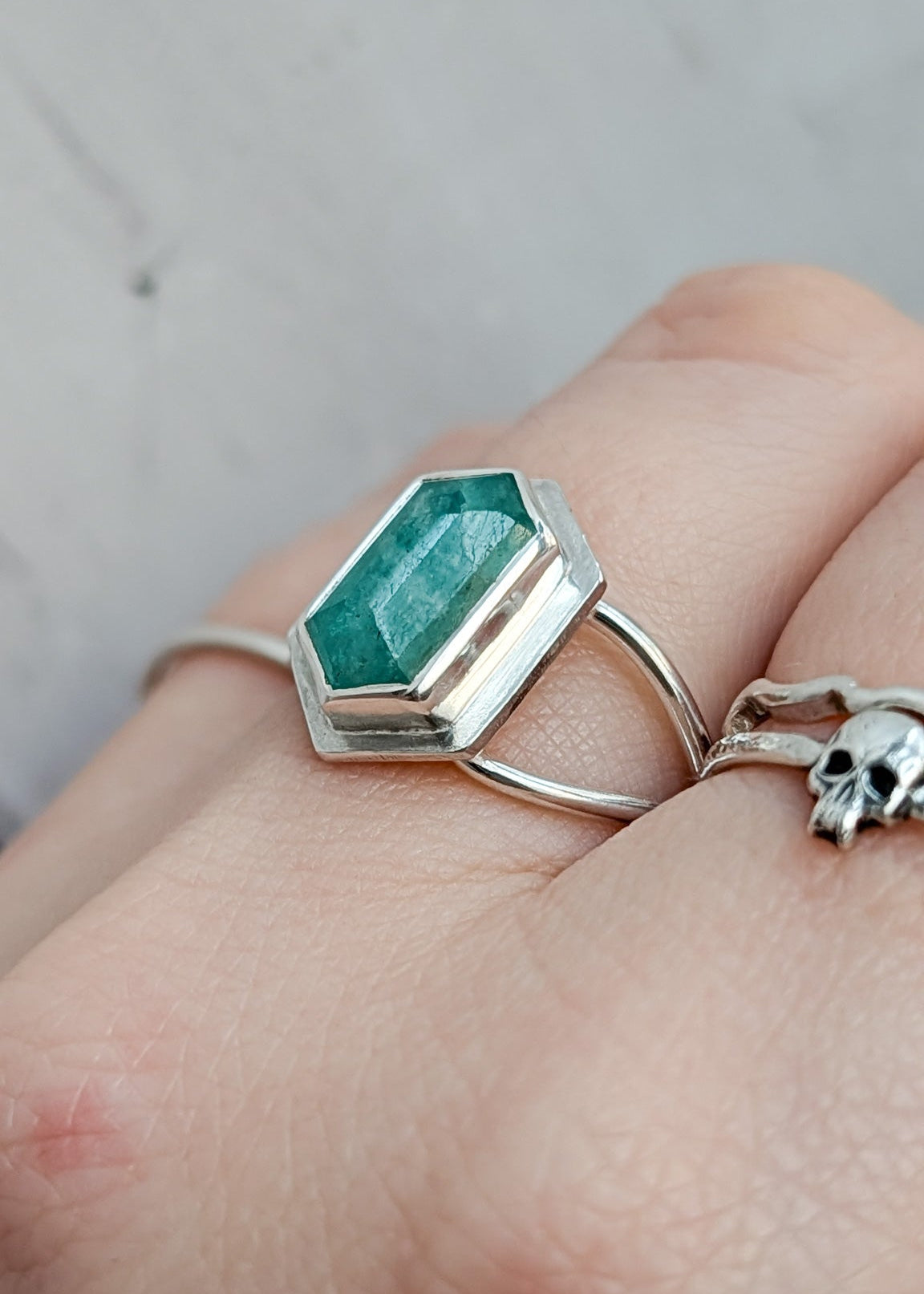 A silver ring with a faceted green gemstone, shown worn on a person's finger alongside a silver skull ring.