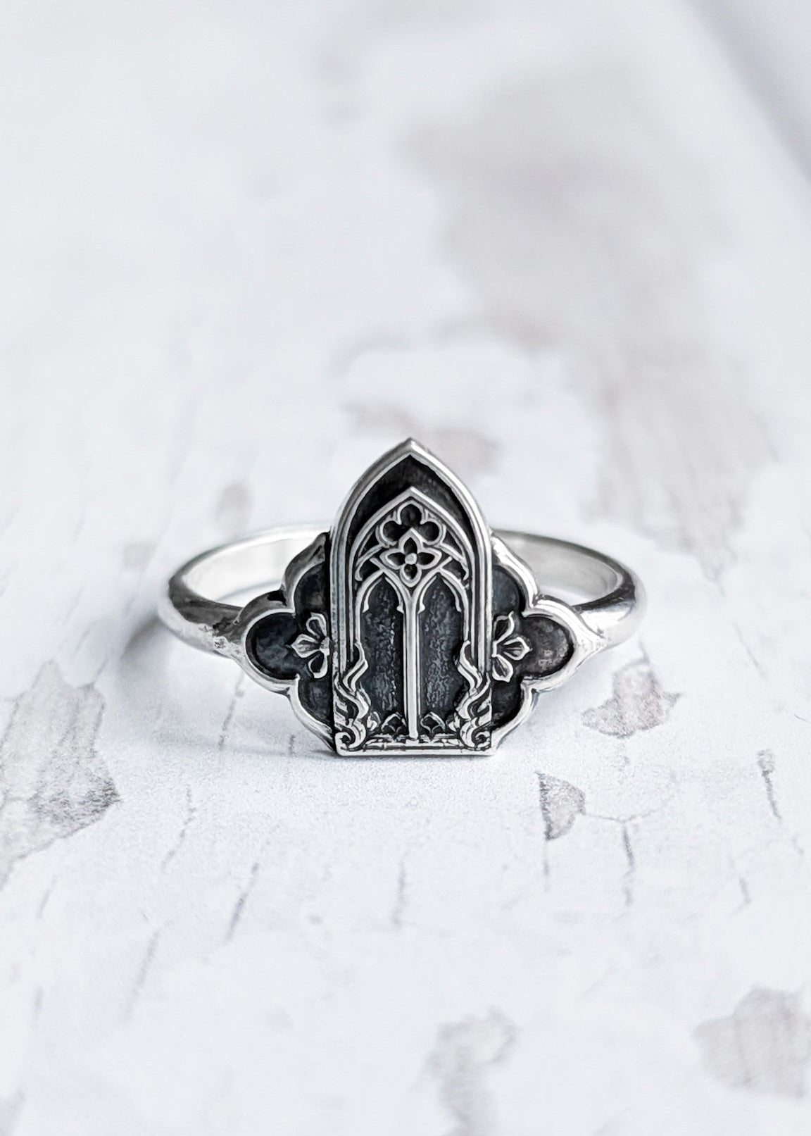 A symmetrical gothic architecture saddle ring with darkened silver details rests on a textured white wooden surface