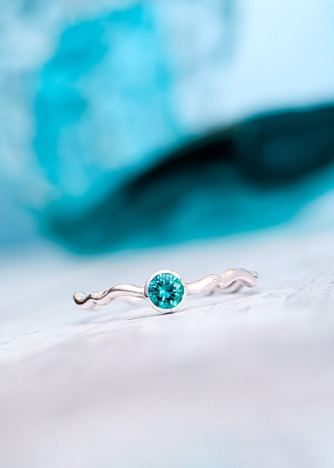 Silver crimped ring with a blue green gemstone resting on a white slanted surface in front of a soft-focus, blue ocean wave