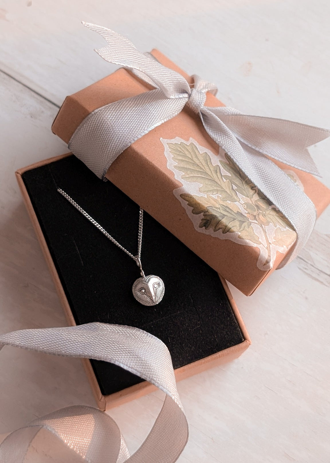 Silver barn owl pendant necklace in a pretty neutral box with a white ribbon on a light wooden surface.