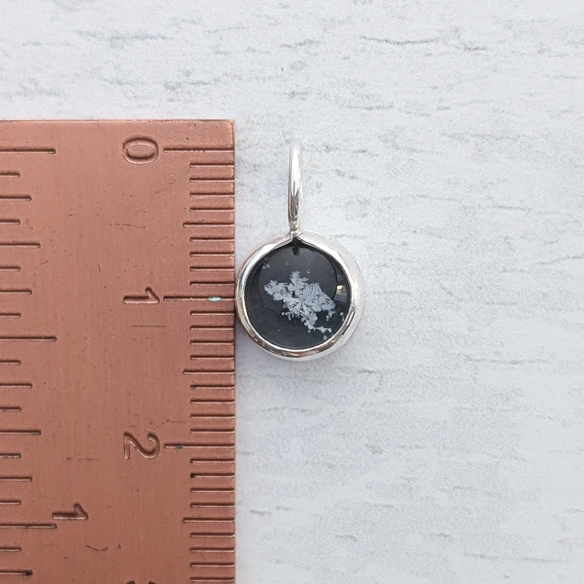 Black and silver real snowflake pendant charm on a light wooden surface beside a ruler for scale.