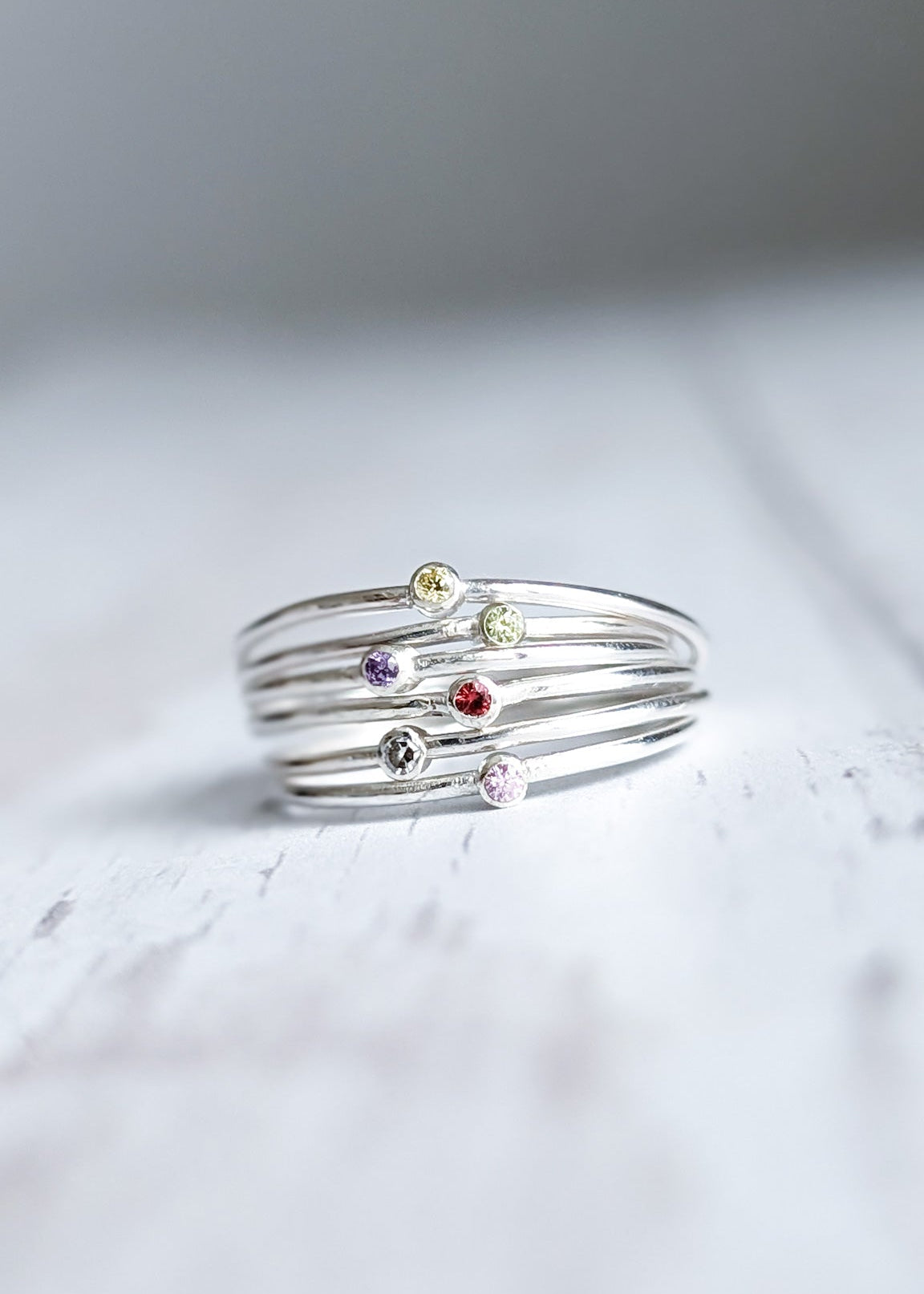 A stack of skinny silver band stacking rings, each with one central stone, all a different colour. Pictured on a white wooden surface.