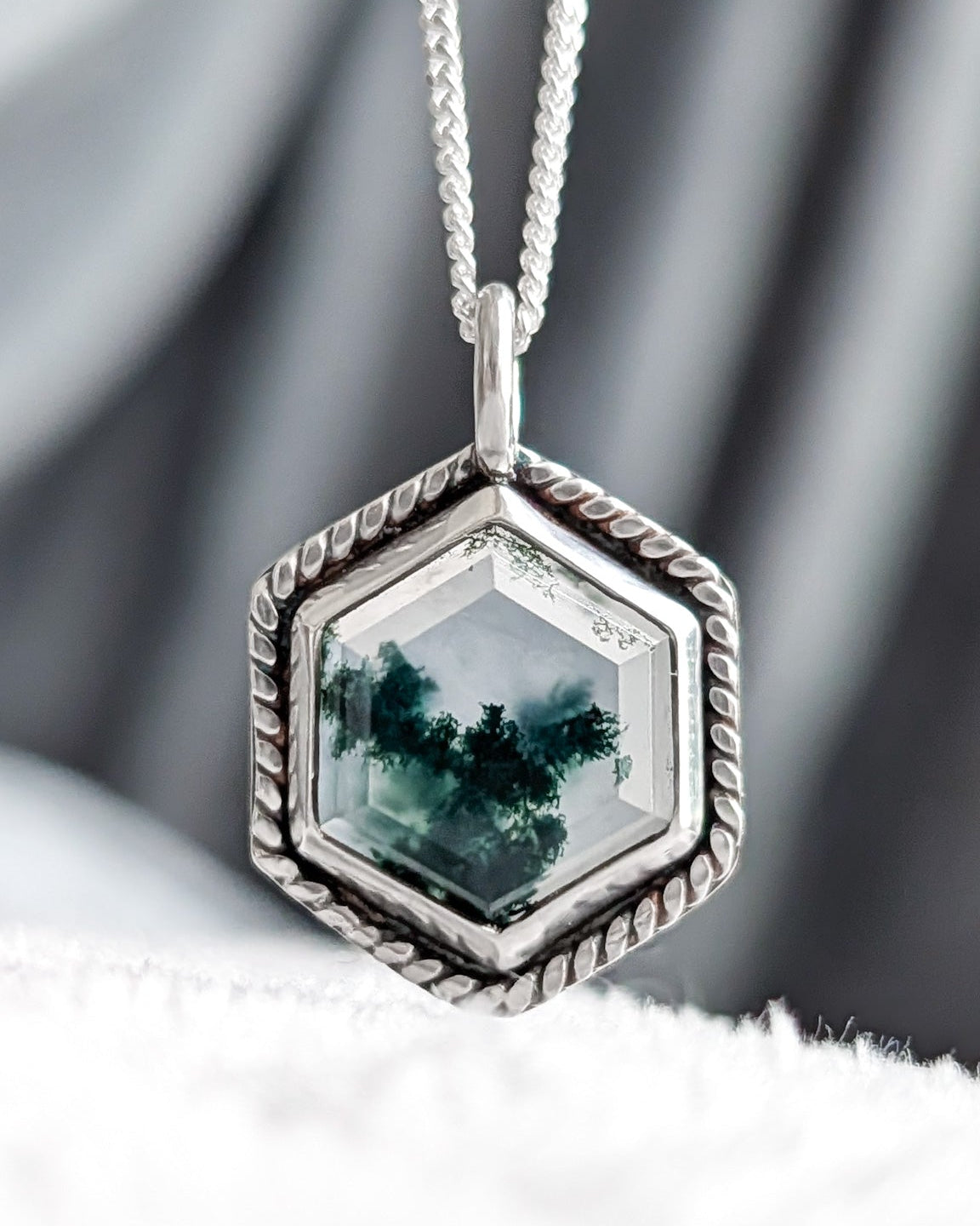 A silver hexagonal pendant necklace featuring a green forest scene gemstone, resting on a white textured surface with a gray draped fabric background.